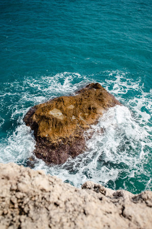 A dramatic view of the sea with vibrant blue water and white foam swirling around a prominent,...