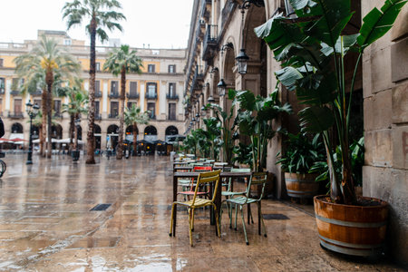 Empty outdoor cafe seating area in a European city square with palm trees and arcades, reflecting...