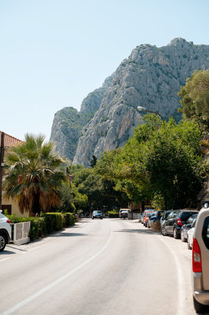 A paved road winds through a coastal town in Croatia, flanked by lush greenery, parked vehicles, and imposing rocky mountains under a clear blue skyの写真素材