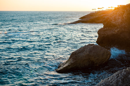 The sun casts a warm glow on the tranquil ocean waters and rugged shore, creating a serene coastal...