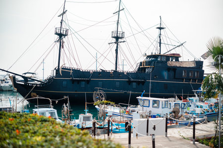 A large, dark-hulled sailing ship with multiple masts and rigging is moored in a busy marina, surrounded by various smaller boats and yachts