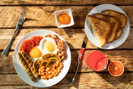 Traditional morning English breakfast - scrambled eggs, white beans, tomatoes, mushrooms, sausages, and apricot jam with classic England fried toast on a white plate and red-orange juice - top viewの写真素材