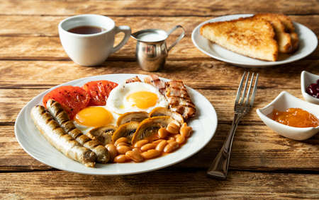 Traditional morning breakfast in England - sausages, tomatoes, eggs, bacon, mushrooms, white beans, toasted toast, a cup of black coffee and jam on a wooden table - side view and close-upの写真素材