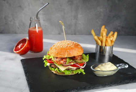 Traditional classic burger consisting of meat, sauce, green lettuce, bacon, tomatoes, and cucumbers with red orange juice with an iron straw, and French fries on a black serving board - close-upの写真素材