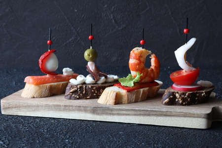 A set of traditional Spanish tapas sandwiches with seafood - octopus, squid, salmon, quail egg, radish, green salad, and pepper on a board on a black backgroundの写真素材