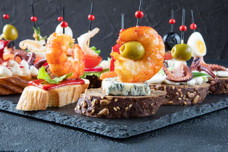 Traditional Spanish food - tapas of a large fried shrimp on gorgonzola cheese with a green olive in the middle on a black baguette with sesame seeds - side viewの写真素材