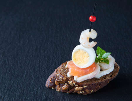 Creative traditional food - Spanish tapas on a black serving board. Tapas are made with seafood - salmon, shrimp, squid, quail egg, and basil leafの写真素材