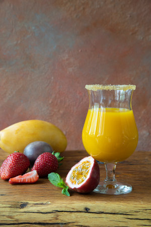 A refreshing summer drink of yellow-orange sunny color in a glass with a border of sugar cane. A cocktail consists of mango, strawberry, mint, passion fruit on a wooden table in the rays of the sunの写真素材