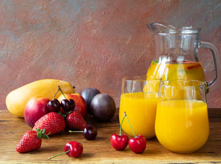 Summer yellow drink made of tropical fruits on a beautiful wooden table with a background of strawberries, cherries, mango, nectarine, and passion fruit and a glass jug with a refreshing cocktailの写真素材