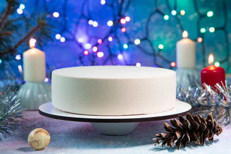 Christmas cake made of French mousse surrounded by garlands, burning candles, a ball on the Christmas tree, pine branches and cones and tinsel on a frosty blue backgroundの写真素材
