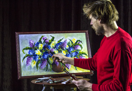 A man bearded artist in a red sweater draws an artistic brush painting flowers still life on easer in the studioの写真素材