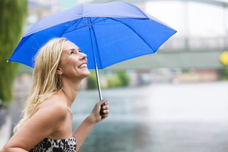 blond woman with an umbrella standing by a river and enjoying the rainy weatherの写真素材