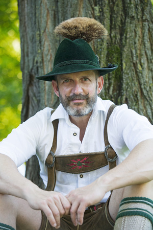 handsome man in traditional bavarian clothes sitting by a treeの写真素材