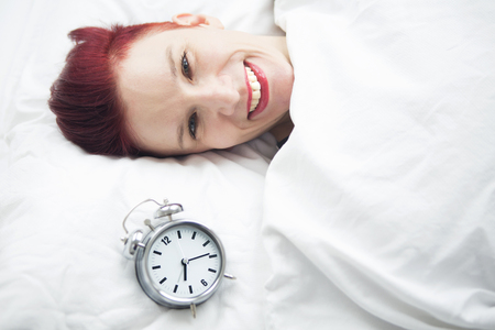 smiling red-haired woman sleeping in bed with alarm clock next to herの写真素材