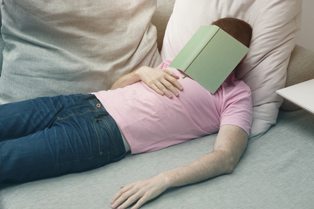 man in jeans and tshirt sleeping on couch with book on his headの写真素材