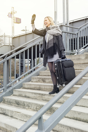 blond woman with suitcase at train station walking down stairs and greeting someoneの写真素材