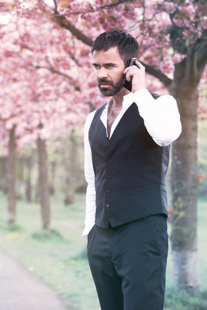 businessman standing in park and talking on phone in front of cherry blossomの写真素材