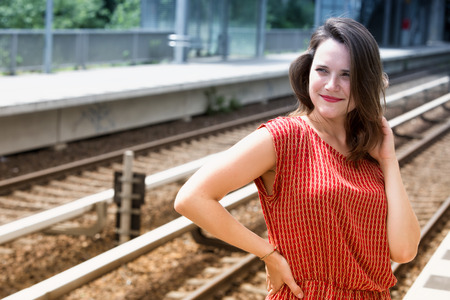 young woman in red dress waiting at platform of train station and smilesの写真素材
