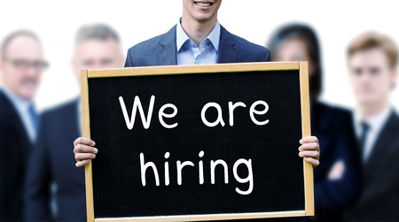 young businessman holding a sign with the words We Are Hiring and group of people behind him