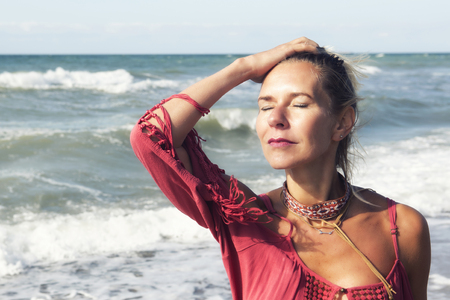 blond woman in red dress standing by the ocean enjoying the sunの写真素材