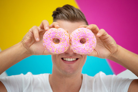 handsome blond man holding two pink donuts in front of colorful backgroundの写真素材