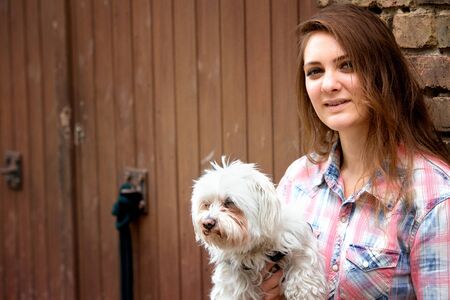 young brunette woman holding her little dog and standing at brick wall