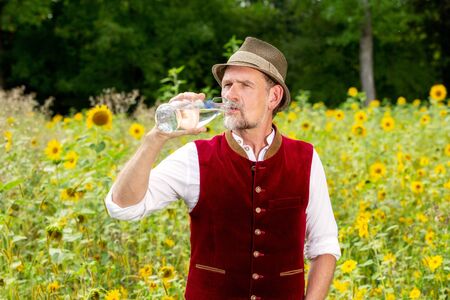handsome bavarian man standing in field of sunflowers and drinking water from a bottleの写真素材