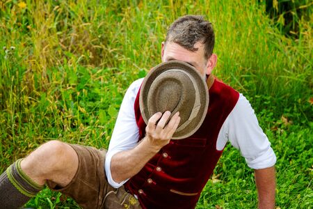 portrait of bavarian man sitting on grass and hiding behind his hatの写真素材