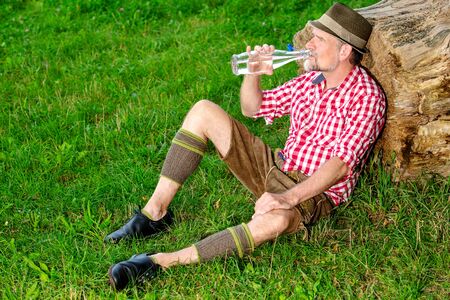 portrait of handsome bavarian man sitting outdoors at tree stump and drinking waterの写真素材