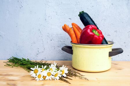 bell pepper, carrots and corgette in rustic pot on wooden table with daisies on grey backgroundの写真素材