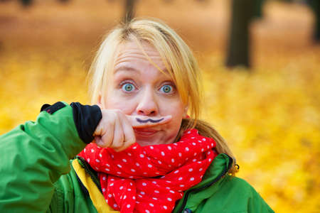funny portrait of young woman in park in autumn with mustache painted on her fingerの写真素材