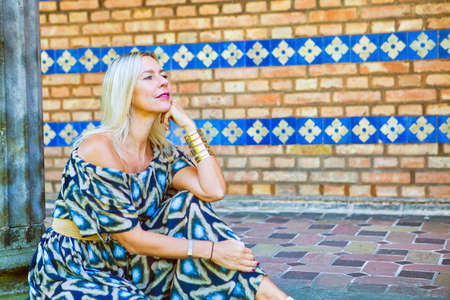 portrait of blond woman in colorful dress sitting outdoors by a pillarの写真素材