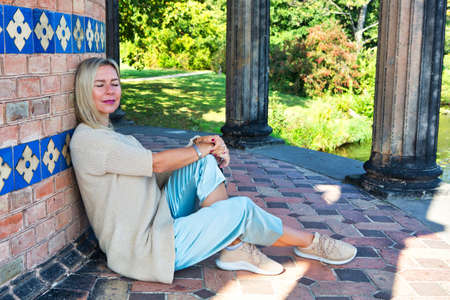 blond woman sitting against a wall by a lake and enjoying the sunの写真素材