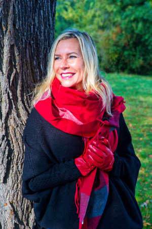 portrait of blond woman with red scarf and gloves standing next to a treeの写真素材