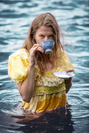 blond woman in yellow historic dress standing in the water holding a teacupの写真素材