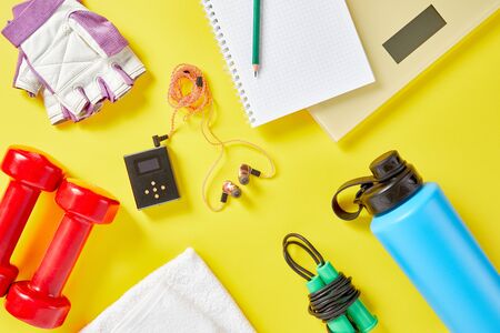 Healthy lifestyle, lose weight and sport concept. Athlete's equipment: red dumbbell, sport water bottles, blank notebook, jump rope, sport gloves, ear phones on yellow background.の写真素材