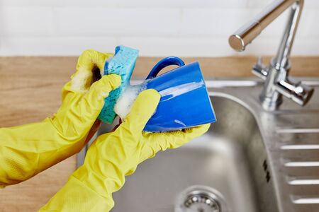 Hands with sponge wash the cup under water, housewife woman in yellow rubber protective gloves washing blue mug in a kitchen sink with a blue sponge, Hand cleaning dishwasher.close up.の写真素材