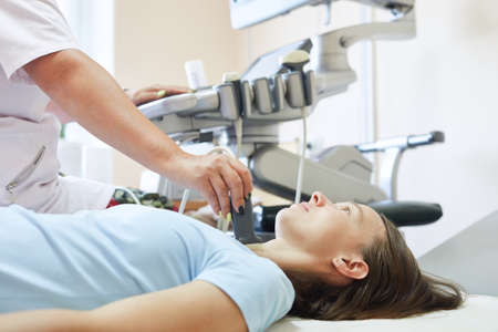 Woman patient during the ultrasound examination of a thyroid lying on the couch in medical office. Girl having neck ultrasound scanning test. Sonographer on background.の写真素材