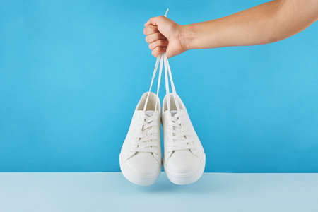 The hand holds by shoelaces Pair of fashion stylish white sneakers on a pastel blue background.の写真素材