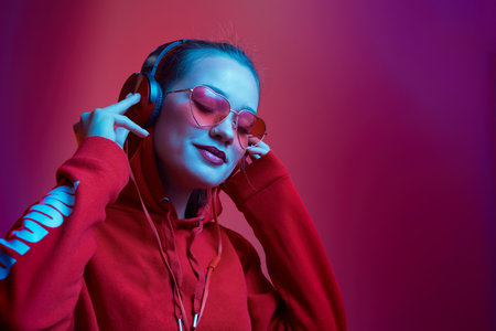 Fashion hipster woman wear stylish glasses and headphones listening to music over color neon background at the studio.の写真素材