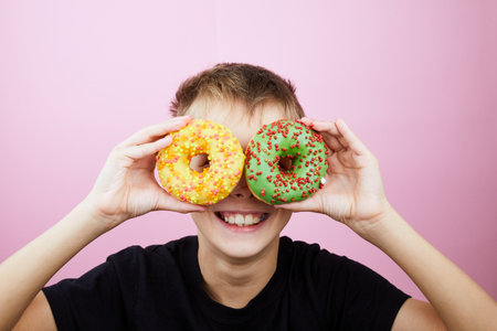 Happy boy is having fun played with donuts on pink background. Child holds donut glasses. Close up.の写真素材