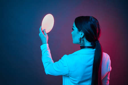 Attractive woman with long hair and white jacket in neon light. Girl looking in mirror.の写真素材