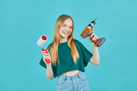 Beautiful girl with an electric screwdriver and an paint roller on blue background. Instruments accessories for renovation apartment room. Repair home concept.の写真素材