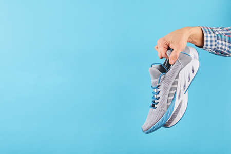 Hand holds hanging gray running sneakers on blue pastel background. Hand with a new sport shoe....