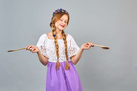 Cheerful little chef girl with hair braids in apron holds a wooden spoon on a gray background....