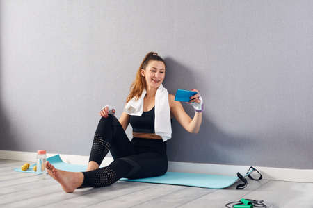 Portrait of fitness woman learning new exercises watching online workout tutorials on her phone at home. Young girl doing fitness in her apartment.の写真素材