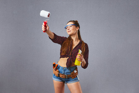Young longhaired woman makes renovation in gloves with paint roller on gray background. Attractive...