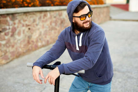 Bearded stylish man riding the electric scooter. Guy on daily commute riding micro scooter. Male in...