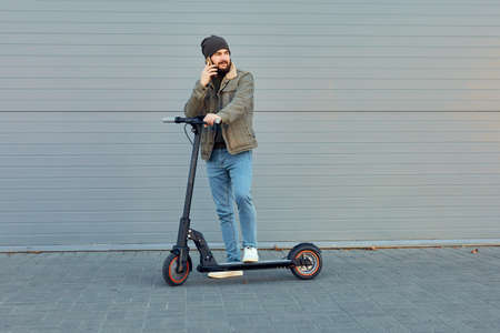 Man with electric scooter near wall. Ecological transportation concept. Stylish bearded male using phone, posing on e-scooter.の写真素材
