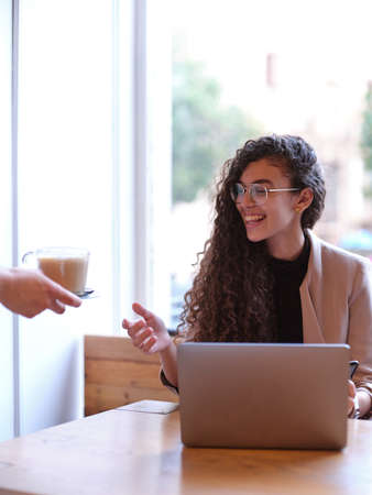 smiling woman receiving a coffee from the waiter while working on her computerの写真素材